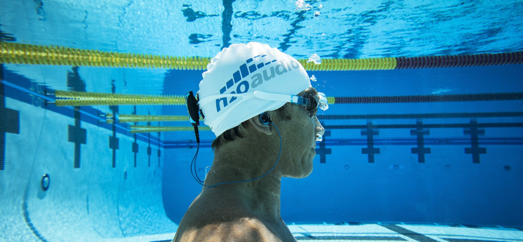 A man in a swim cap is submerged in a swimming pool while listening to music from the Stream waterproof MP3 player made by H2O Audio. 