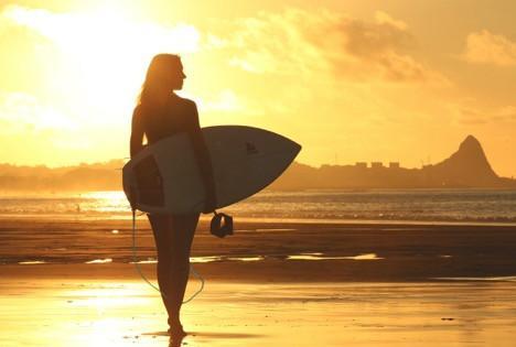 An attractive, athletic woman walks toward the setting sun while holding a surfboard. 