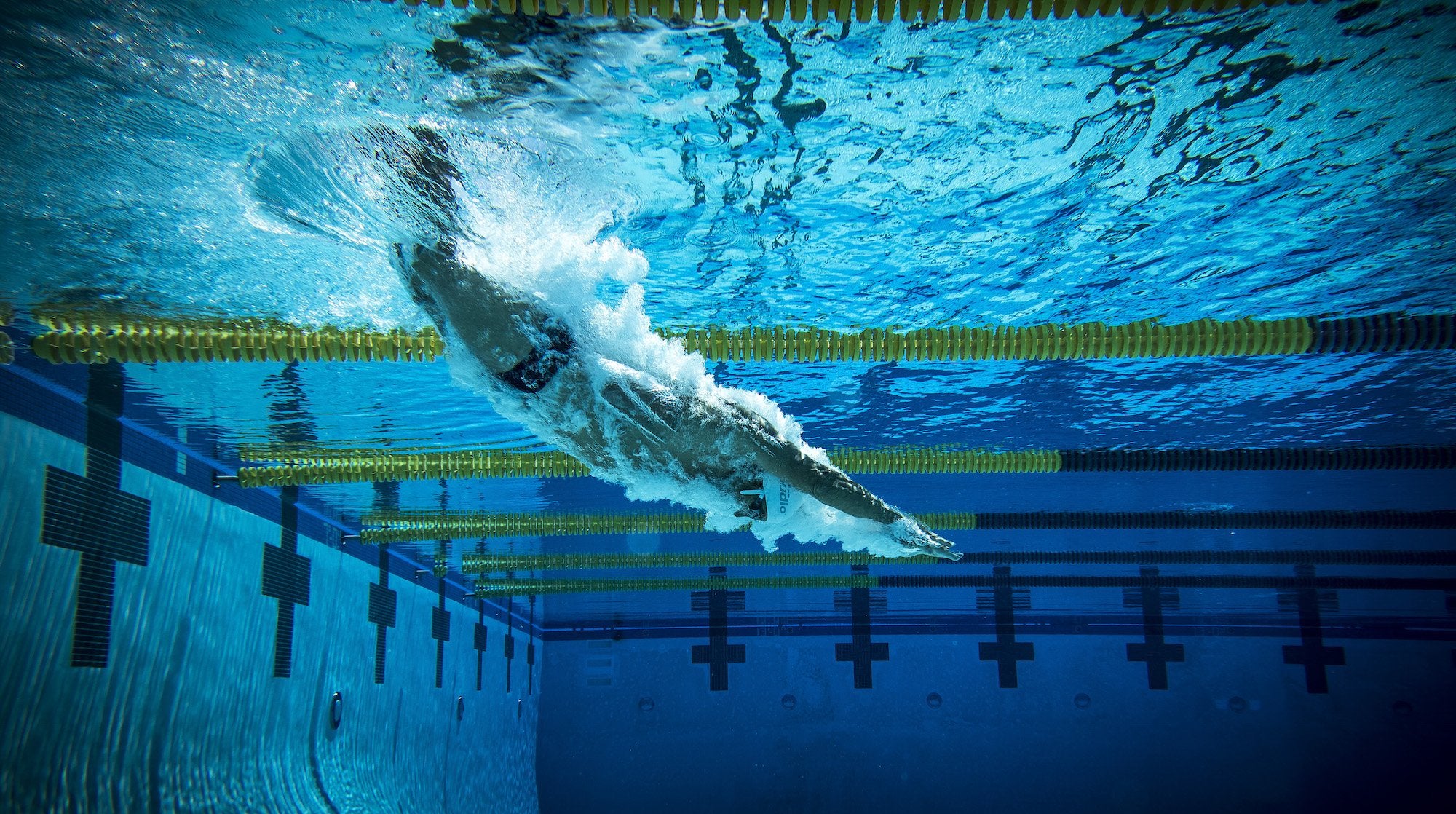 A male swimmer dives under the water while wearing custom waterproof headphones made by H2O Audio. Listening to music helps many swimmers to stay motivated during work outs weight-loss and relaxation.