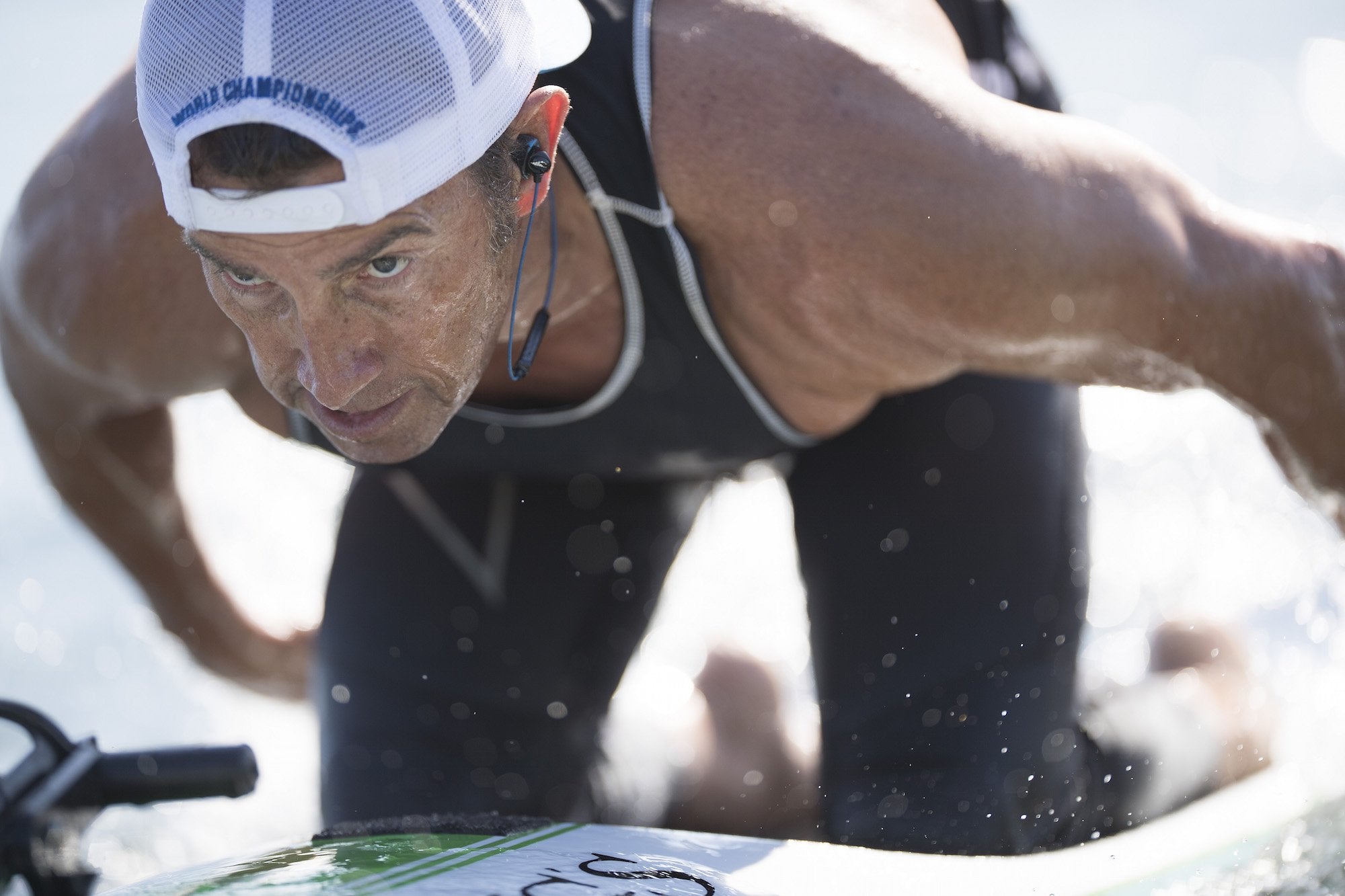 A male focuses ahead while paddleboarding on his knees. Waterproof headphones and music players (or waterproof phone cases) make it possible for ocean athletes to listen to music while working out, without the risk of damaging their devices.