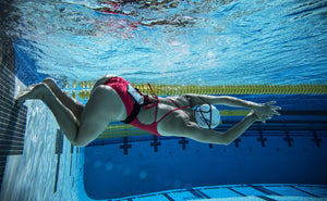 A female swimmer wearing a red one-piece swimsuit pushes off the wall of a swimming pool while listening to music with H2O Audio waterproof headphones and the Amphibx case for smart phones. 
