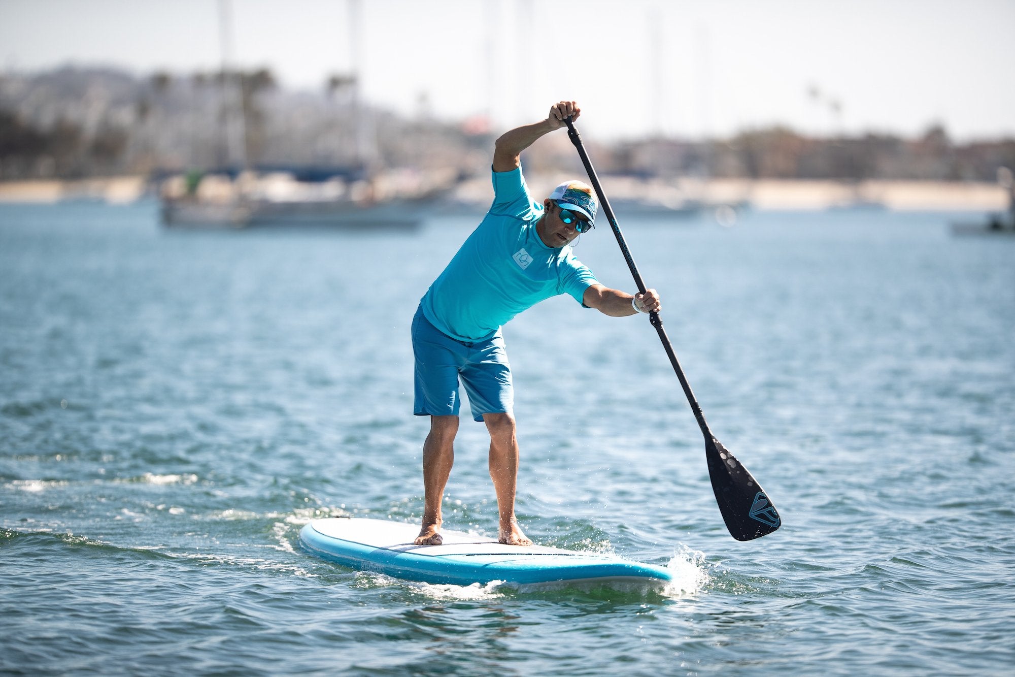 An athletic man wearing blue shorts stand-up paddleboards in a harbor while listening to music and using waterproof headphones from H2O Audio. 